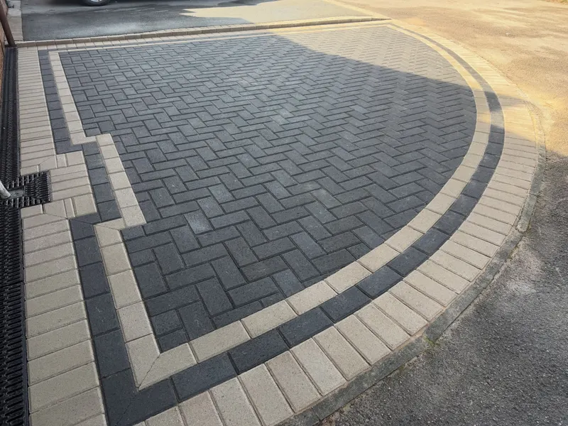 Charcoal herringbone block paving with alternating buff and dark border stripes, slot drain and cut-outs around wall fittings.