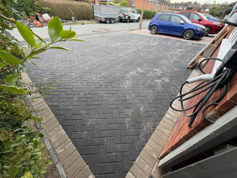 Charcoal herringbone block driveway with a tan soldier border at the road edge, red brick house and parked cars nearby.