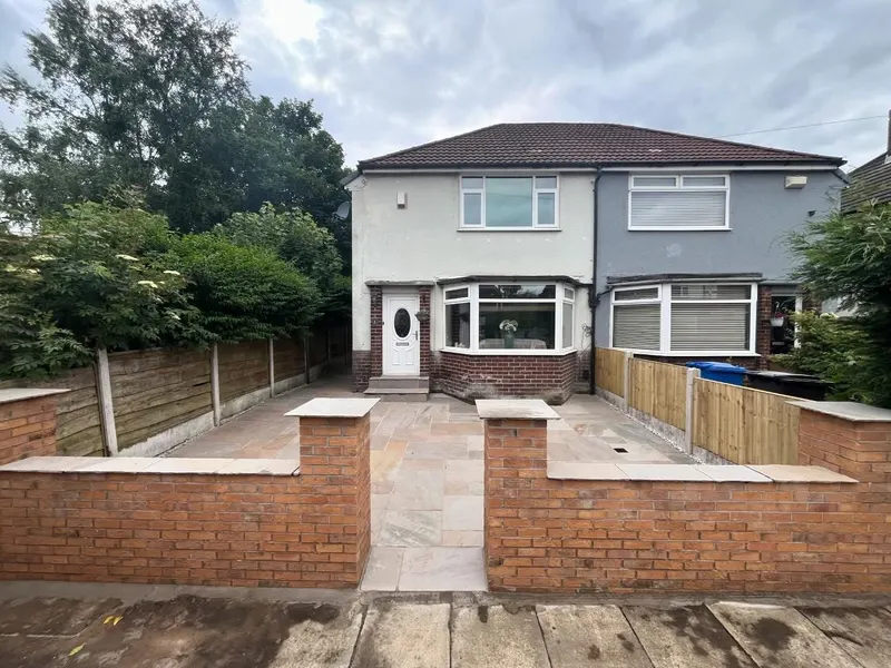 Red brick perimeter wall with grey stone capping and a central opening, fronting a large buff and grey natural stone paved front garden.