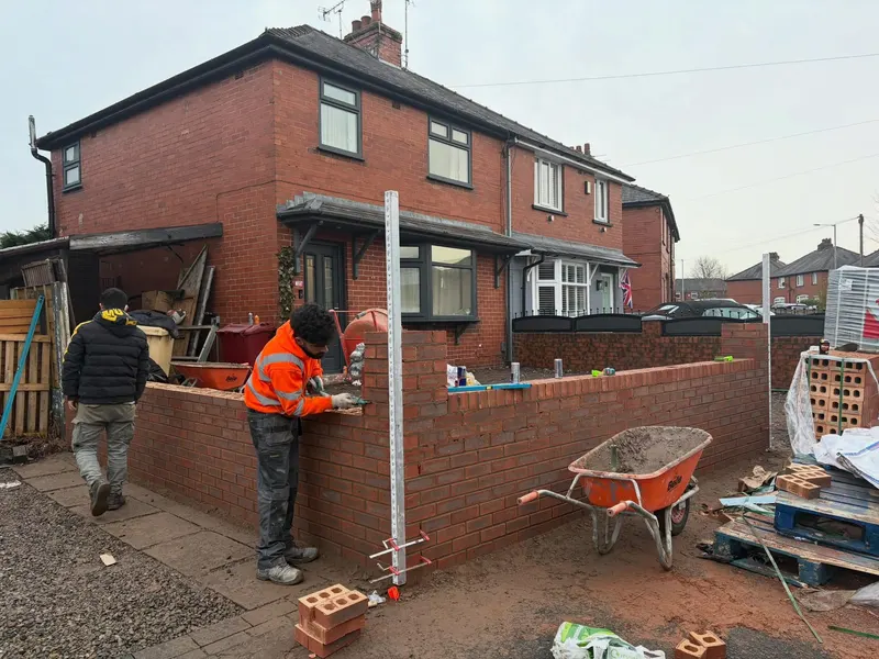 Brickwork in progress as workers build a waist-high red brick boundary wall on a front garden site, with mortar, bricks, and tools nearby.