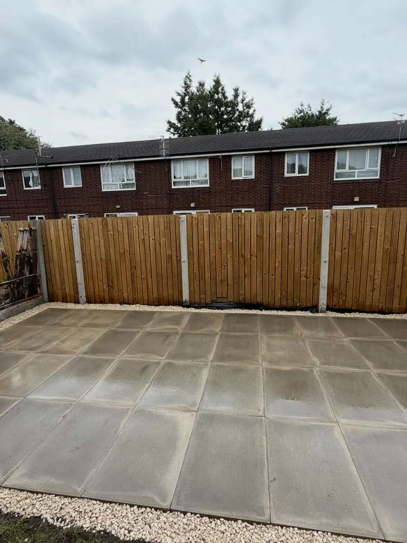 New vertical timber slat fencing on concrete posts, seen beyond a grid of light-grey patio slabs and a narrow gravel strip.
