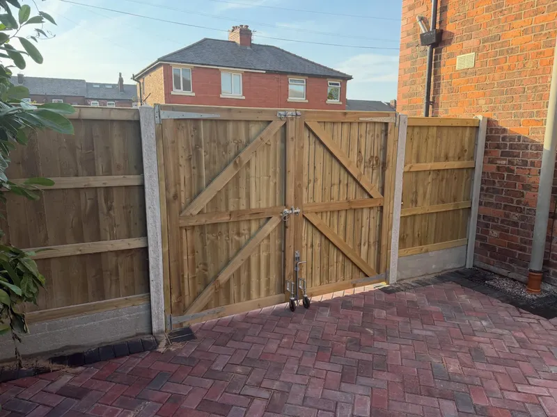 Pair of braced wooden driveway gates with metal hinges and drop bolts, flanked by featheredge fence panels on concrete posts above herringbone block paving.