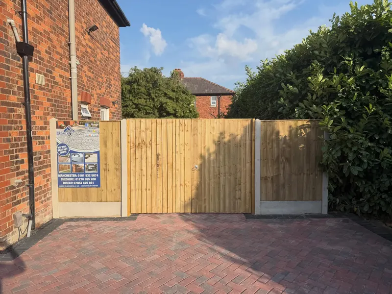Double vertical-board wooden gates and matching fence panels between a brick house and a hedge, set at the end of a herringbone block-paved driveway.