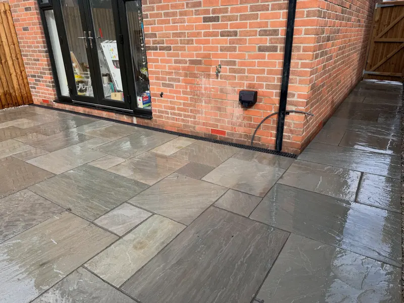 Multi-tonal sandstone flags laid beside black-framed French doors on a red brick house, with a matching side path and a linear drain at the wall.