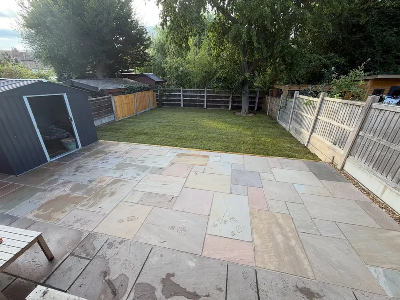 Expansive mixed-tone natural stone patio beside a green lawn, with a metal shed on one side and timber panel fencing around the garden.