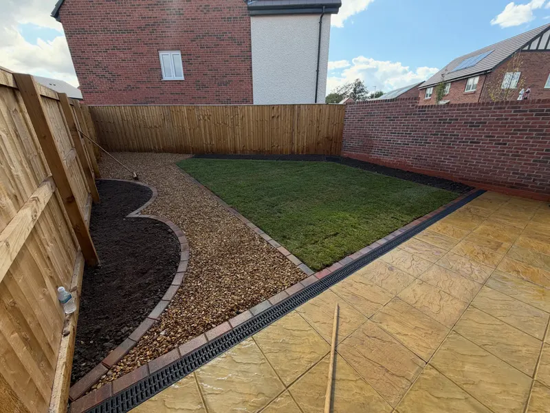 Square buff-toned riven paving slabs beside a fresh turf lawn, gravel paths, and curved brick-edged planting beds in a fenced rear garden.