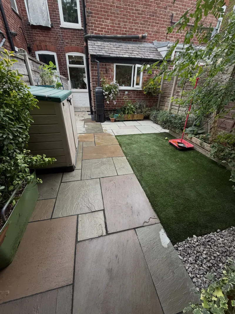 Irregular natural stone slabs forming a walkway and patio around a rectangular artificial grass patch, with gravel and brick walls in a compact yard.