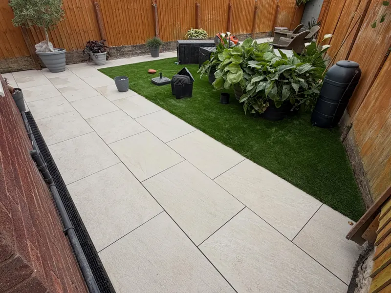 Large light beige rectangular patio tiles in a running-bond layout beside bright artificial turf, with a channel drain along a brick wall and potted plants.