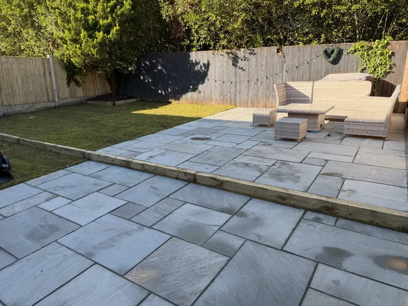 Light grey mixed-size stone flags on a rear patio with rattan seating, timber sleeper edging, lawn to one side, and painted wooden fencing.