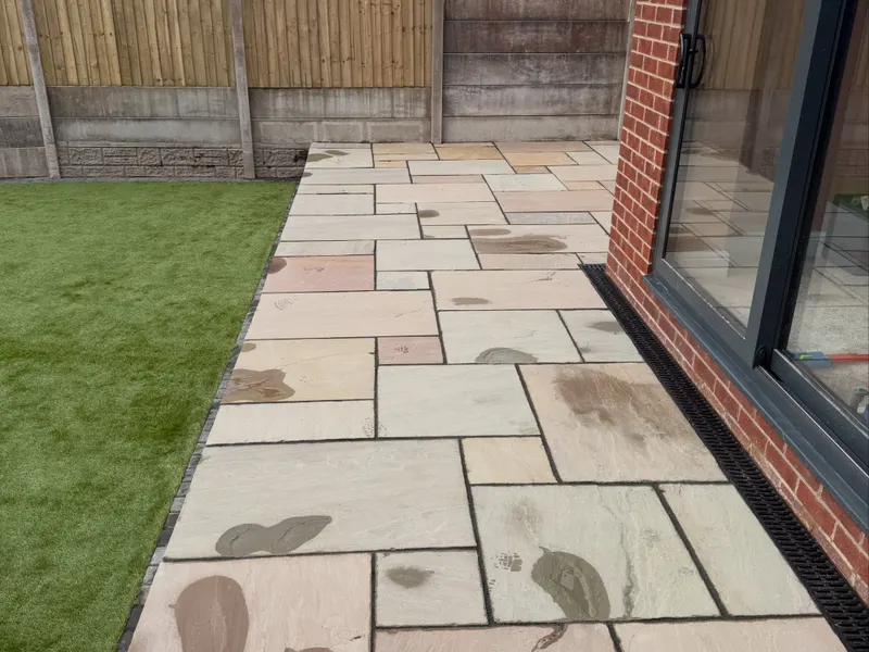 Damp multi-tonal stone flags running along a red brick wall with dark-framed sliding doors, block edging, and a slot drain at the base.
