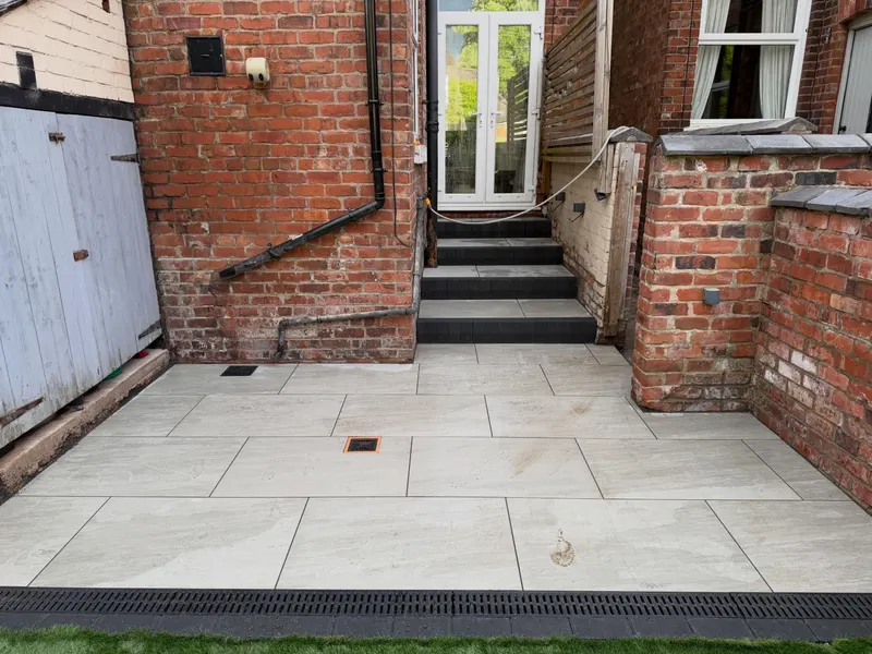 Large light grey paving slabs with contrasting dark risers on steps up to white French doors, channel drainage, and brick walls on three sides.