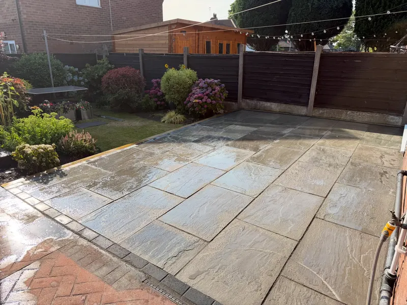 Wet light grey stone flags bordered by charcoal block pavers, meeting an older herringbone brick area, with lawn, shrubs, and wooden fencing beyond.
