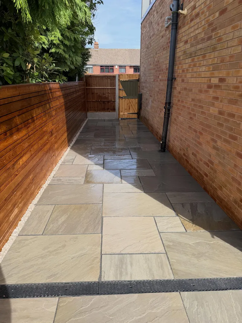 Mixed sandstone flags along a narrow side return between a brick house wall and a horizontal timber fence, with gravel edging and a gate at the end.