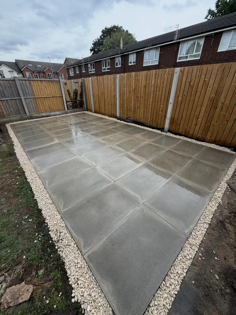 Smooth light grey square paving slabs in a grid pattern, surrounded by a decorative gravel border and new vertical timber fencing.