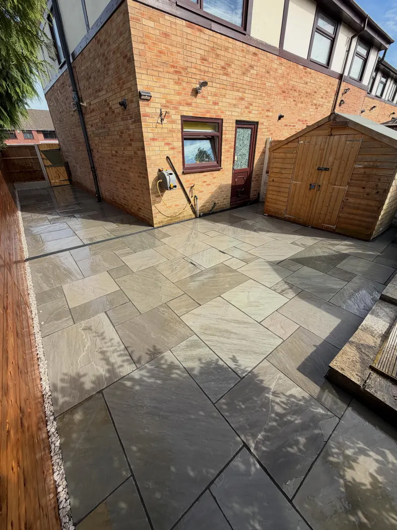 Multi-size natural stone flags covering a rear patio and side path beside a brick house, with white gravel edging, a channel drain, and a wooden shed.