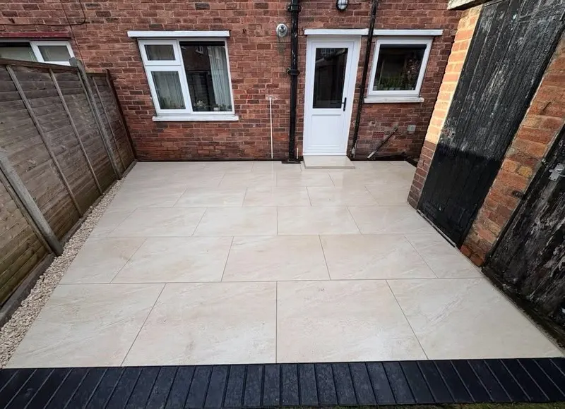 Large cream rectangular patio tiles in stretcher bond beside red brick walls, with a dark ribbed border, gravel edging, and a wooden fence.