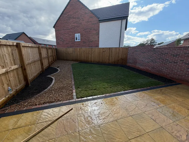 Buff riven square stone slabs with a slot drain and dark block edging, surrounding a central turf lawn and gravel beds in a fenced garden.
