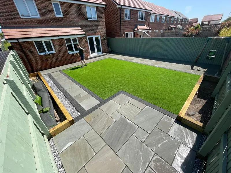Light grey mixed-size stone flags with a charcoal block border around a rectangular artificial lawn, timber sleeper beds, and a painted fence.