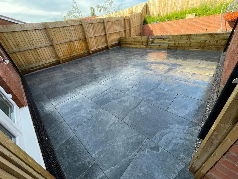 Large dark grey textured patio tiles beside a brick house, with a timber sleeper retaining wall, steps, gravel edging, and a channel drain.
