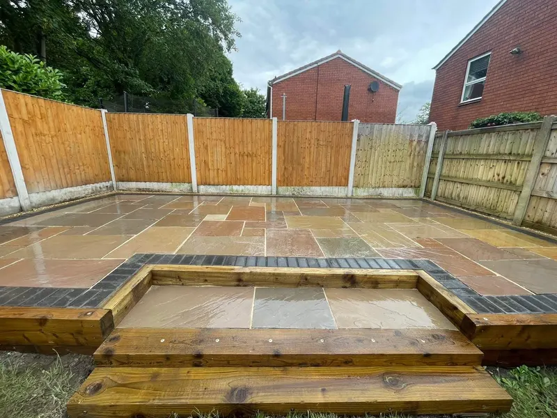 Multi-tonal natural stone patio framed by dark block edging, with timber sleeper steps and tiers at the front of a fenced garden.