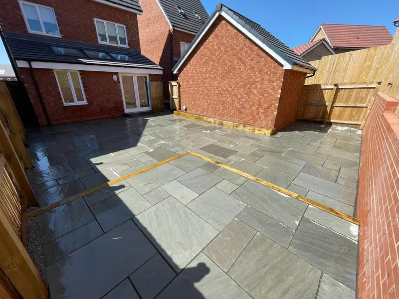 Blue-grey natural stone flags laid between red brick house walls and outbuildings, divided by timber sleeper borders under a clear sky.
