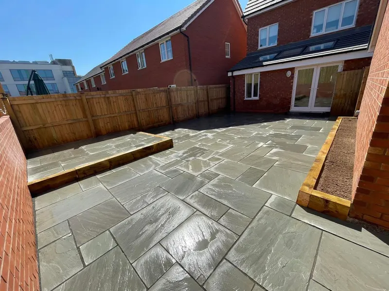 Mid-grey riven stone flags with a timber sleeper raised section and a long sleeper planter bed filled with soil beside a brick extension.