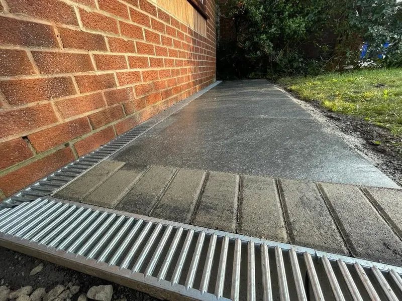 Dark grey textured paving slabs with light block borders and metal slot drains running alongside a red brick wall toward a lawn.