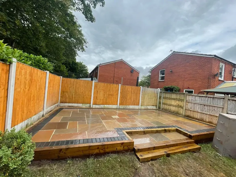 Elevated multi-tonal stone patio within timber sleeper walls and dark block edging, with sleeper steps down to bare ground in a fenced yard.