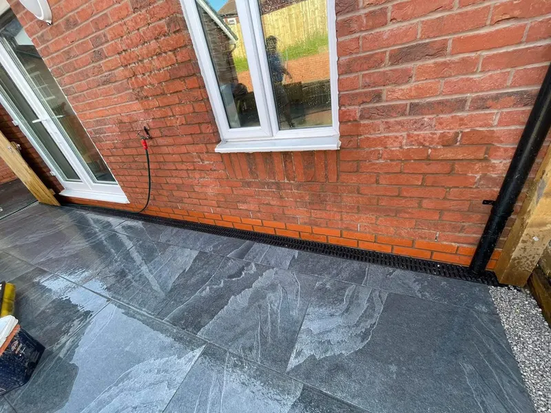 Large dark grey veined slabs laid tight to a red brick wall with a channel drain, white door and window, and gravel at one edge.