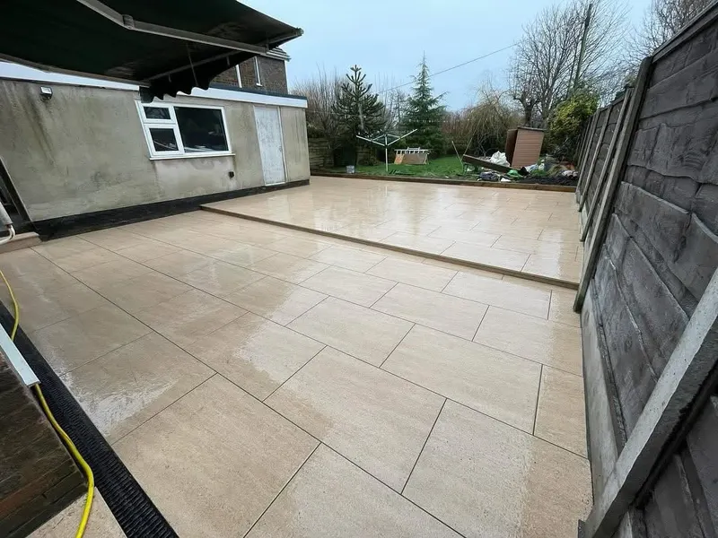 Light beige large-format paving on lower and upper tiers with a single step, beside a rendered wall, wooden fence, lawn, and trees.