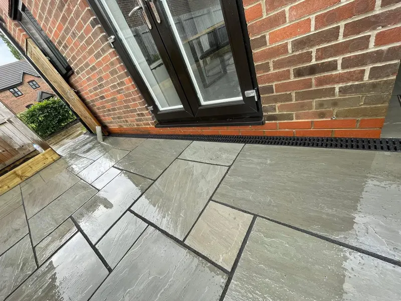 Wet light grey riven stone flags with dark pointing along a brick house, meeting dark-framed French doors and a wall channel drain.