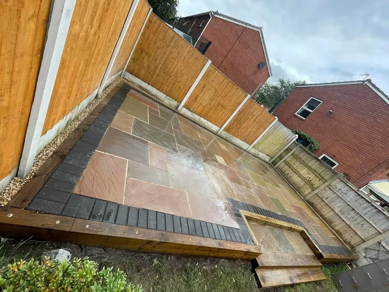 Wet multi-tonal stone flags on a raised platform edged with charcoal blocks and timber sleepers, with gravel beside a new fence.