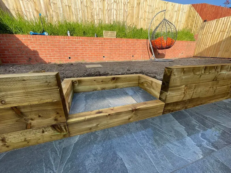 Blue-grey large-format paving at the base of a stepped timber sleeper retaining wall, with exposed soil above and a brick boundary wall.