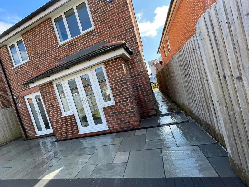 Reflective grey mixed-size stone flags behind a brick house with bi-fold doors, continuing down a side alley with a channel drain and dark border.