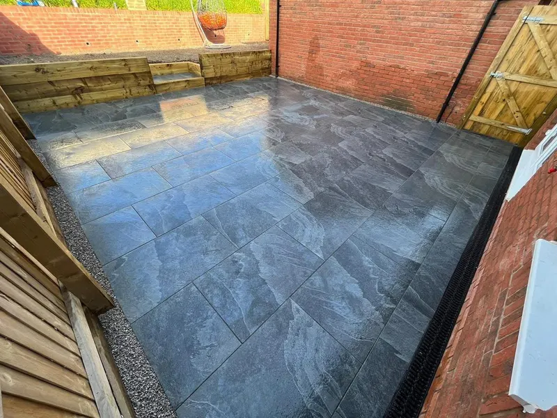 Charcoal grey veined rectangular slabs in stretcher bond within brick and timber-fenced walls, with sleeper retaining tiers and gravel edging.