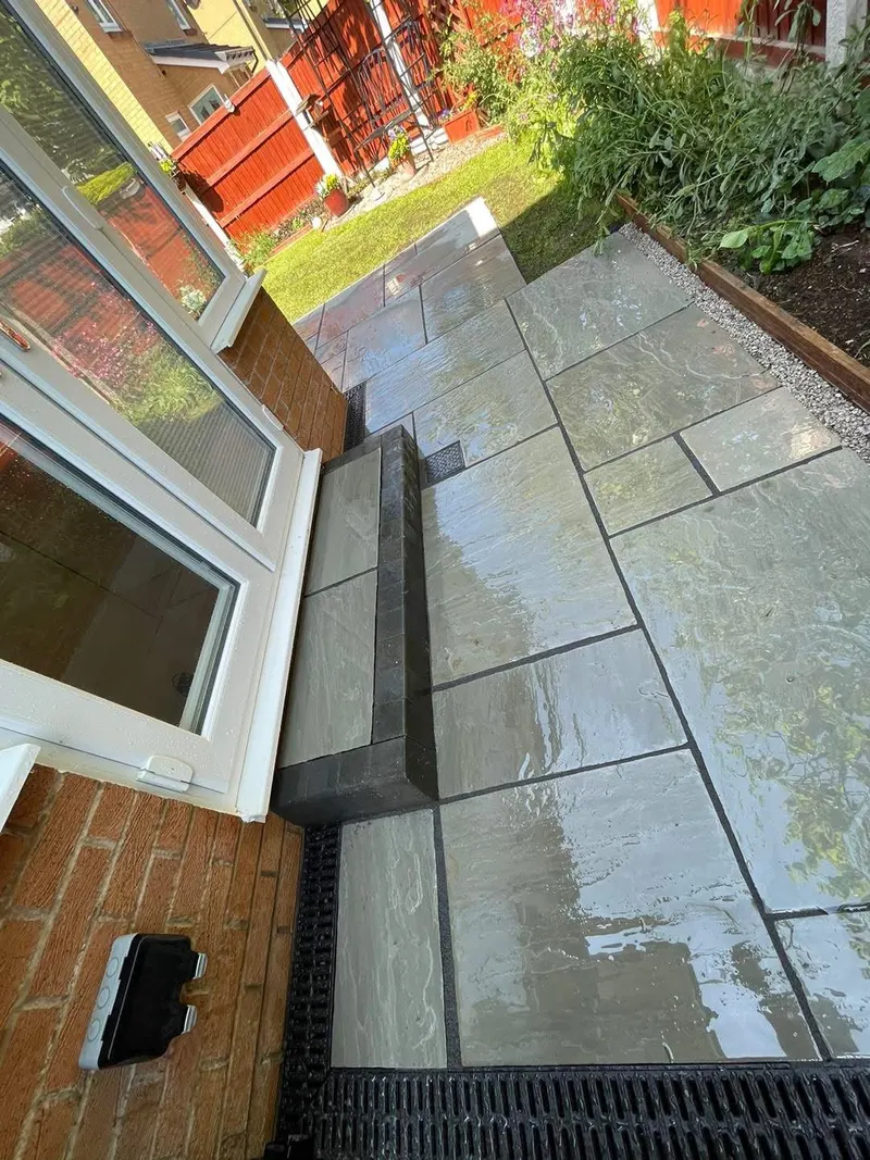 Wet multi-toned natural stone patio with a single paved step edged in dark blocks, channel drains, gravel, and a timber-edged planting bed.