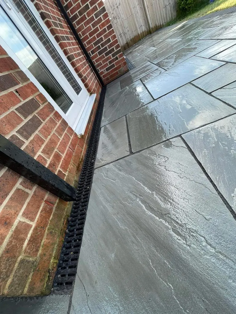 Large grey riven stone slabs in random bond beside a brick wall and window, with a black channel drain running along the foot of the wall.