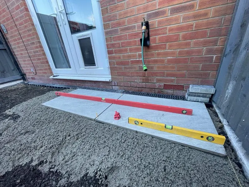 Two light grey patio slabs set on aggregate beside white patio doors, with spirit levels, a channel drain, and excavated ground still visible.