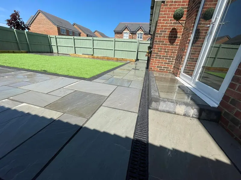 Light grey and buff stone flags with a dark block border around artificial grass, a paved step to the house, and a painted fence.