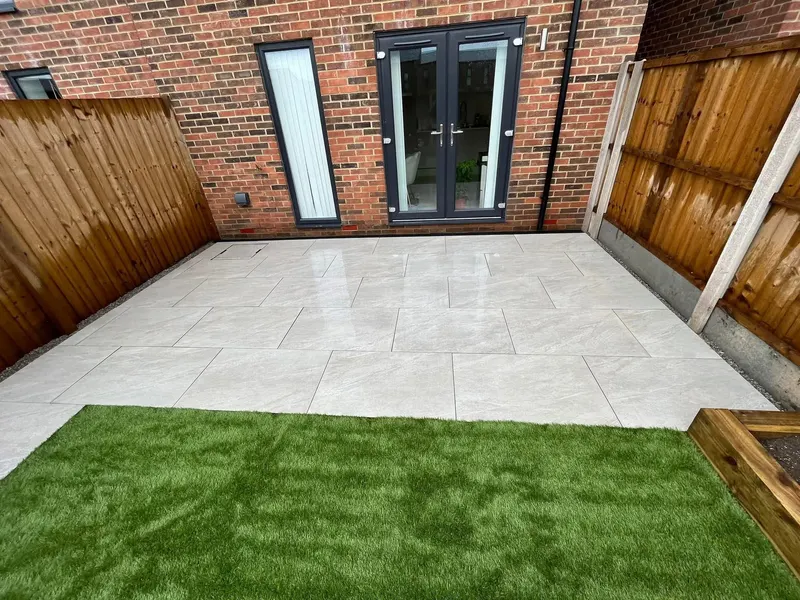 Pale grey veined porcelain-style slabs in stretcher bond against a red brick house with charcoal French doors, artificial grass, and timber fencing.