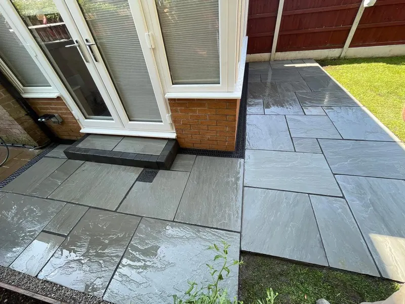 Grey natural stone flags with a matching paved step and dark block risers, slot drains along brick and white conservatory walls, beside lawn.