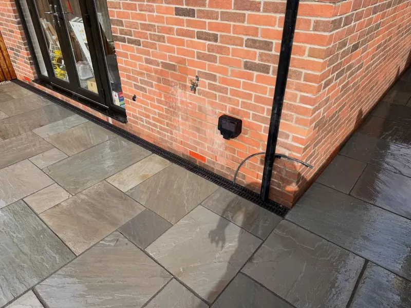 Close view of grey, buff, and tan riven stone slabs meeting a brick wall, black-framed doors, and a flush channel drain.