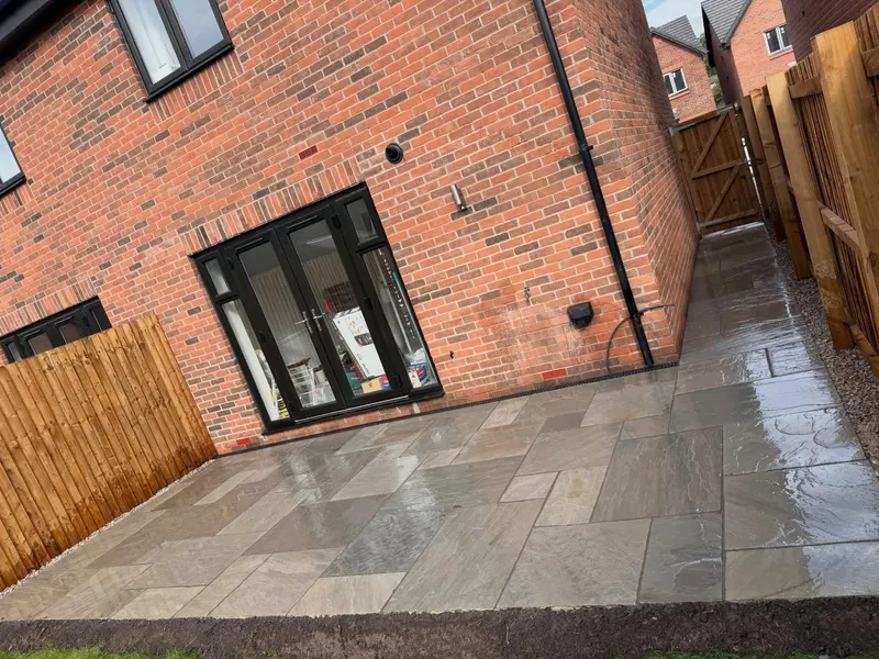 Wet mixed grey and buff stone flags outside glass doors, continuing as a side path with gravel edging toward a wooden gate.