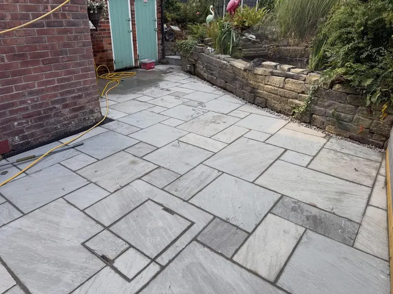 Light grey modular stone patio with an inset cover and channel drain, bordered by gravel and a rough stone retaining wall with planting.