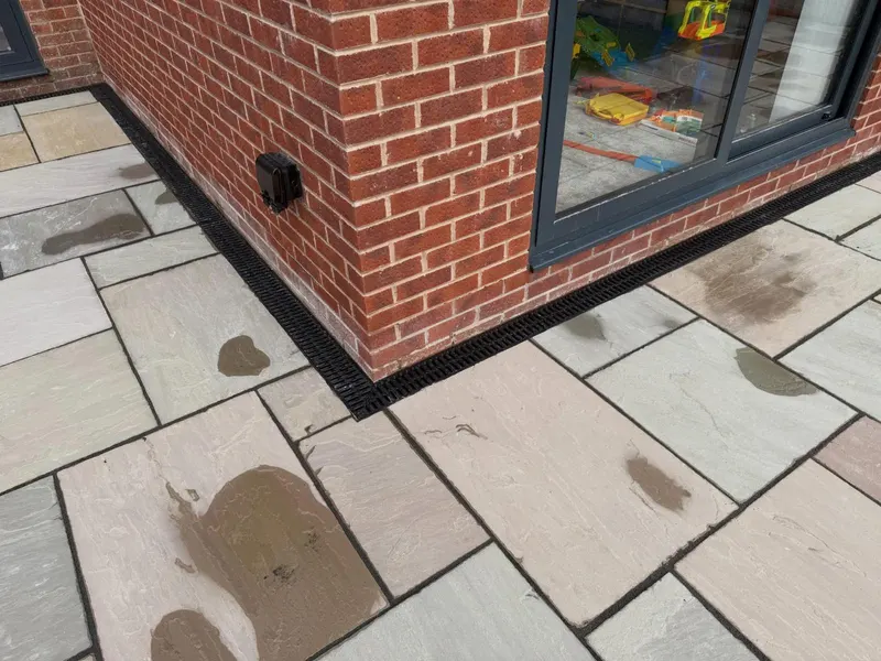 Multi-tonal riven stone slabs at an external brick corner with dark pointing, wet patches, and a channel drain along the wall.