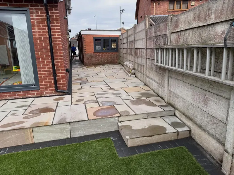 Natural stone flags on upper and lower levels with dark block edging and a wide step, beside a brick extension and side path to an outbuilding.