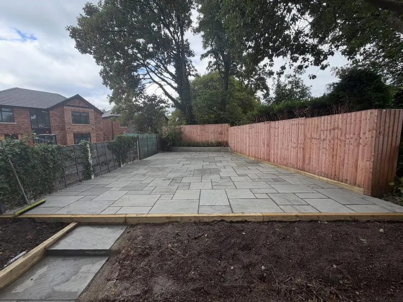 Light grey rectangular stone slabs in a mixed layout, framed by timber sleepers and fencing, with neighbouring houses visible behind.