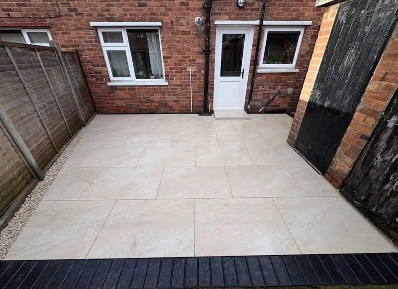 Large cream textured patio tiles from a brick house to an outbuilding door, with gravel beside a vertical fence and a dark front border.