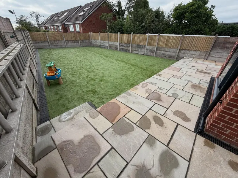 Buff and grey natural stone flags around a rectangular artificial grass area, with a channel drain at the brick wall and block edging on the fence side.