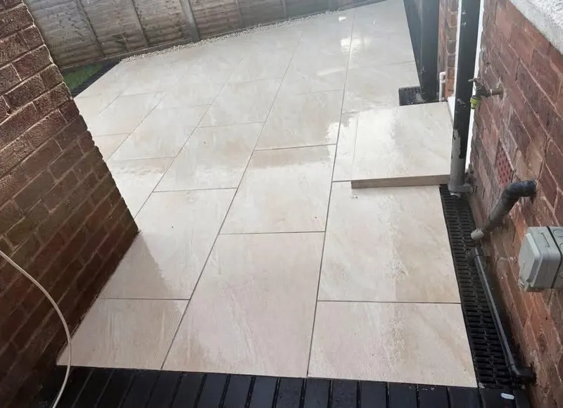 Light beige large-format slabs in stretcher bond between red brick walls, with a channel drain, small paved step, and gate at the end.